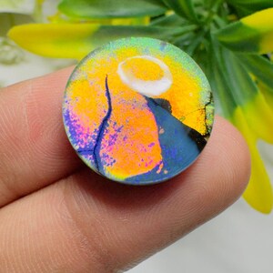 May include: A round, dichroic glass pendant with a vibrant design. The pendant features a yellow and orange background with a white crescent shape and a blue and purple mountain-like shape. The edges have a cracked appearance.