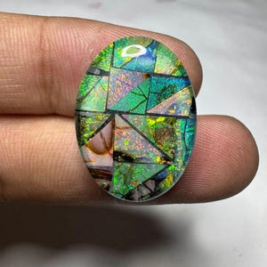 May include: An oval-shaped doublet gemstone featuring a mosaic pattern of iridescent green, blue, and orange fragments set against a dark background. The surface displays a high-gloss finish with distinct geometric segments separated by dark lines.
