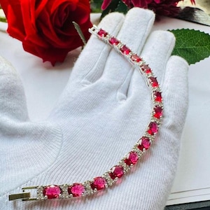 May include: A silver-toned tennis bracelet featuring a series of oval-cut vibrant red stones alternating with small clear accent stones. The bracelet has a secure metal clasp closure and is displayed against a white fabric background.