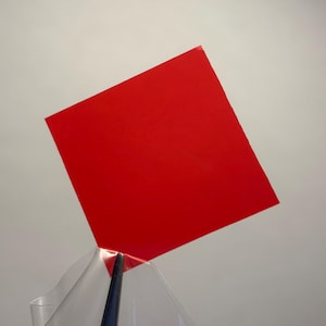 May include: A vibrant, solid red square-shaped piece of material held by a pair of dark-coloured tweezers. The square is positioned at an angle, displaying a smooth, flat surface with sharp, clean edges and a uniform, bold red colour throughout.