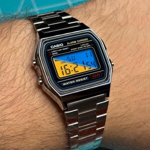 May include: A silver-tone digital watch with a metallic link band. The rectangular watch face features a black border, a blue and yellow digital display showing the time 16:21, the text CASIO, ALARM CHRONO, LITHIUM, and WATER RESIST.