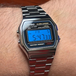 May include: A silver-tone digital watch with a stainless steel link band. The rectangular face features a blue backlit display showing the time 5:47:33 and the day FR 5. Text on the watch includes CASIO, ALARM CHRONO, LITHIUM, WATER RESIST, and WR.