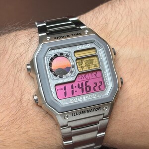 May include: A silver-tone digital wristwatch with a stainless steel link band. The rectangular face features a world map display, a circular analog-style clock graphic with a sunset landscape, and a pink digital screen showing 11:46:22 PM.