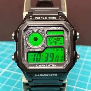 Puede incluir: Un reloj digital Casio negro con una pantalla verde vibrante. La esfera presenta un mapa mundial, un dial analógico circular y una lectura de hora digital que muestra las 10:39. El texto incluye WORLD TIME, WR100M, 10 YEAR BATTERY e ILLUMINATOR.