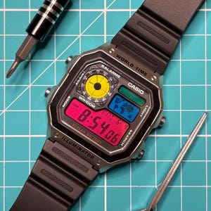 May include: A Casio digital watch with a black resin strap and a silver-tone case. The face features a yellow circular analogue-style display, a blue world map graphic, and a red digital screen showing 8:54:06, FRI 6-4. Text includes WORLD TIME, CASIO, WR100M, and ILLUMINATOR.