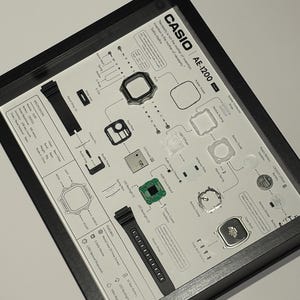 May include: A framed exploded view display of a Casio AE-1200 watch, featuring individual components like the circuit board, LCD panel, battery, and resin straps arranged on a white background with technical labels and specifications.