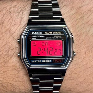 May include: A silver-tone digital wristwatch with a metallic link band. The rectangular watch face features a bright red digital display showing the time, day, and date. Text on the face includes CASIO, ALARM CHRONO, Lithium, and WATER RESIST.