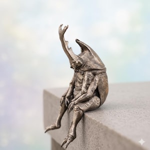 Puede incluir: Una escultura de bronce de una criatura parecida a un escarabajo sentada en un borde. La escultura presenta detalles intrincados, incluyendo un cuerno grande y segmentos corporales complejos. La criatura está posada con la cabeza baja y un brazo levantado.