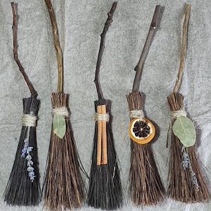 May include: Five small decorative brooms made of dark brown natural bristles and wooden twigs. Each broom is tied with twine and features unique accents: lavender sprigs, a green bay leaf, two cinnamon sticks, or a dried orange slice.