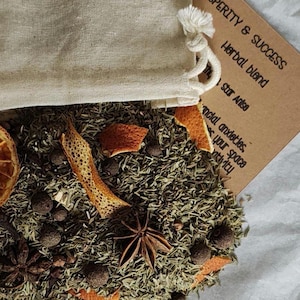 May include: A beige cotton drawstring pouch sits above a pile of loose herbal blend containing dried orange slices, star anise, and dark round spices. A tan rectangular card with the text Prosperity & Success Herbal blend is visible.