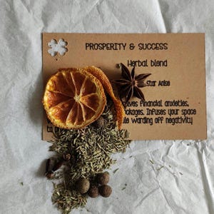 May include: A brown card with the text Prosperity & Success, Herbal blend, Star Anise, and descriptive text. A dried orange slice, a star anise, and a pile of dried herbs and small round dark brown seeds are placed on the card surface.