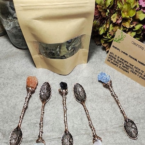 May include: Five antique copper-toned metal spoons with ornate floral embossed bowls and twig-like handles, each topped with a raw gemstone including orange, black, blue, and clear crystals, alongside a brown paper pouch and a card.
