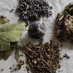 May include: A collection of dried botanical elements including a pile of purple lavender buds, a cluster of green bay leaves, a mound of dried brown herbs, and a rough, translucent purple amethyst crystal positioned in the centre of the arrangement.