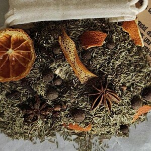 May include: A loose herbal tea blend spilling from a small beige drawstring pouch. The mixture contains dried green herbs, dark brown peppercorns, star anise, and slices of dehydrated orange. A tan paper tag with visible text is attached.