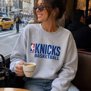 May include: A light grey crewneck sweatshirt featuring the blue and red NBA logo and the text KNICKS BASKETBALL printed in bold blue letters across the chest. The garment has long sleeves with ribbed cuffs and a ribbed neckline, made of a soft fabric.