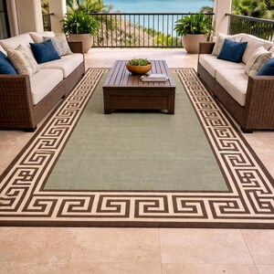 May include: An outdoor patio scene featuring a green rug with a brown Greek key border, a dark brown coffee table, and two wicker sofas with cream cushions and blue pillows. The background shows a view of the sea.