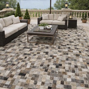 Puede incluir: Escena de patio al aire libre con una gran alfombra hecha de parches cuadrados en tonos grises, marrones y negros. Dos sofás con cojines beige y una mesa de centro están dispuestos bajo una gran sombrilla. El fondo presenta una vista al lago.