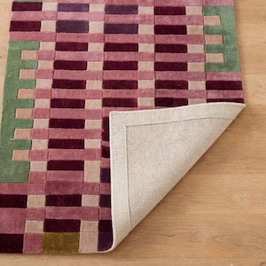 Può includere: Un tappeto rettangolare caratterizzato da un motivo geometrico di rettangoli intrecciati nelle tonalità del viola scuro, magenta, rosa chiaro e verde salvia. Un angolo del tappeto è ripiegato per rivelare un retro in tessuto beige chiaro.
