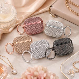 Könnte beinhalten: Sechs mit Strass besetzte AirPod-Hüllen in Roségold, Silber, Gold und Schwarz. Jede Hülle hat einen passenden Ring. Die Hüllen schützen und verleihen Ihren AirPods einen Hauch von Glanz.