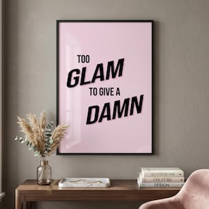 May include: A framed pink poster with the text "TOO GLAM TO GIVE A DAMN" in black. The poster is displayed on a wooden table with a vase of dried flowers and books. The wall is a neutral gray color.