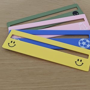 May include: A set of four flat, rectangular plastic rulers arranged in a fan shape. The rulers are yellow, blue, pink, and olive green. The yellow ruler features two black smiley face icons. The blue ruler has a white football graphic. The pink ruler has a small white icon, and the olive green ruler has a black alien head icon.