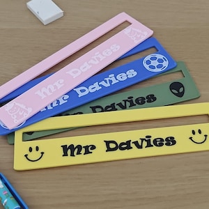 May include: Four rectangular plastic rulers in pink, blue, olive green, and yellow. Each ruler features a horizontal slot and the text Mr Davies printed in a bold font. The pink ruler has a unicorn, the blue has a football, the olive green has an alien head, and the yellow has two smiley faces.