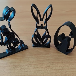May include: Three black Easter-themed cookie cutters. One is a rabbit sitting, another is a standing rabbit, and the third is a rabbit inside an egg shape. The cutters are made of a dark material and sit on a light brown surface.