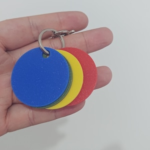 May include: A set of four circular, flat tags in blue, green, yellow, and red, each featuring a small hole at the top. The tags are stacked together and secured by a silver-coloured metal split ring, creating a compact and organised key ring set.