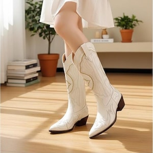 以下が含まれることがあります： 精巧な刺繍が施された白いカウボーイブーツ。 ブーツはポインテッドトゥと茶色のヒールが特徴です。 これらのトールブーツは滑らかな素材で作られており、さまざまな場面に適しています。