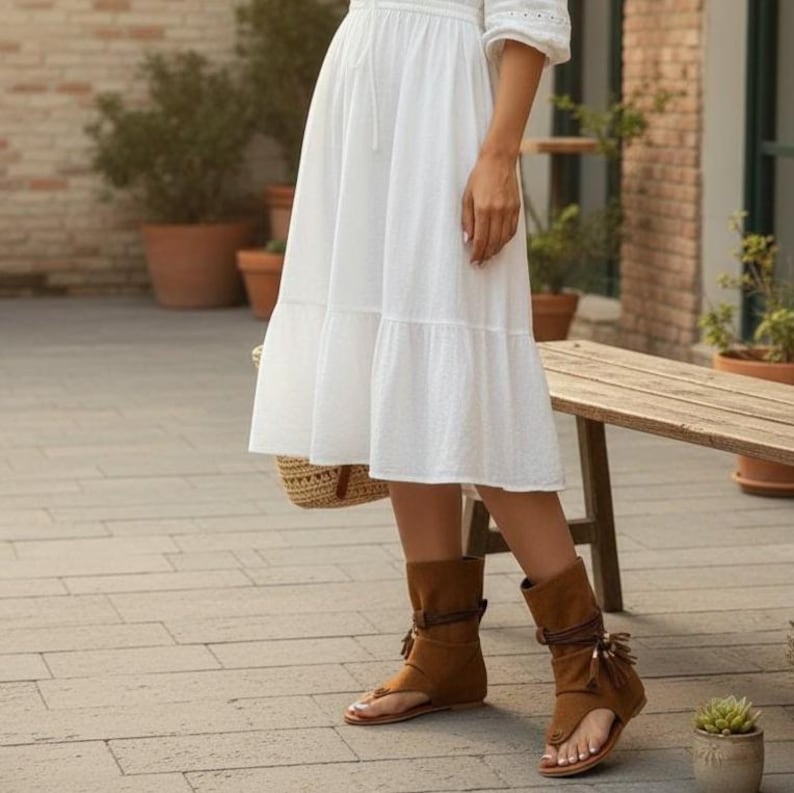 May include: A person wearing a white dress and brown sandals with tassels. The sandals have a thong-style design and are made of suede-like material. A woven basket bag is held in the left hand.