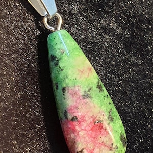 Pode incluir: Um pingente em forma de gota com uma fiança e corrente prateadas. A pedra preciosa é uma mistura de verde, rosa e preto, com uma superfície polida. O pingente é provavelmente uma joia.