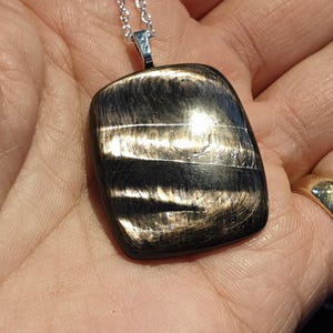 Peut inclure: Un collier avec un pendentif noir et doré et une chaîne argentée. Le pendentif est de forme rectangulaire aux bords arrondis, avec un motif tourbillonnant noir et doré. Une bague dorée est visible sur la main tenant le collier.