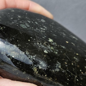 May include: A polished, smooth, oval-shaped stone with a deep black base color. The surface features scattered, small, metallic-looking flecks in shades of silver and gold that catch the light, creating a reflective and glossy appearance throughout.