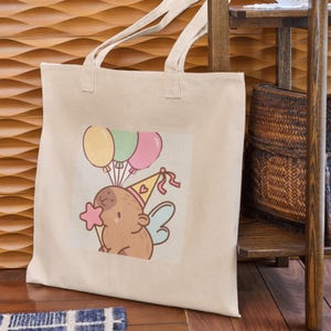May include: Beige canvas tote bag featuring a cartoon illustration of a brown creature wearing a party hat and holding a star wand. The illustration includes three balloons in yellow, green, and pink. The bag has two handles.