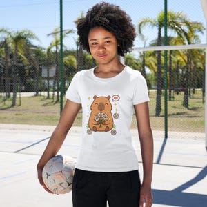 May include: White t-shirt featuring a cartoon capybara holding daisies, with a heart-shaped thought bubble. The capybara is brown with a smiling face. The shirt is worn by a person holding a soccer ball.
