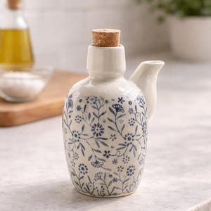 May include: A white ceramic oil cruet with a cork stopper and a small spout. The cruet is decorated with a blue floral pattern. The cruet is designed for dispensing oil and other liquids in the kitchen.