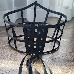 Könnte beinhalten: Schwarzer Kopfschmuck im Käfigstil mit Schnürfront und langen Satinbändern. Der Kopfschmuck besteht aus schwarzem Metall und Stoff und liegt auf einer Holzoberfläche.