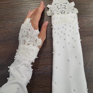 May include: A pair of white fingerless gloves featuring intricate floral lace detailing at the wrist and hand area, accented with scattered white pearls throughout the length of the fabric and a small loop for the middle finger on each glove.
