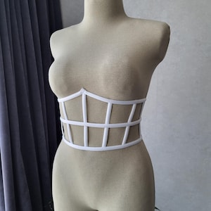 以下が含まれることがあります： マネキンの上に展示された、白いケージスタイルのコルセットトップ。コルセットは、垂直と水平の白いストラップで構成された構造的なデザインで、胴体に格子状のパターンを作り出しています。マネキンはニュートラルなベージュ色です。