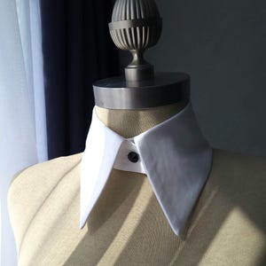 May include: A pristine white detachable collar with a sharp, pointed design, secured by a single black button. The collar is presented on a mannequin, highlighting its tailored form. The background includes a window with natural light.