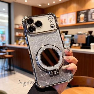May include: Silver glitter phone case with a built-in ring holder. The case has a clear, protective bumper and a reflective ring for a secure grip. The case is designed to fit a smartphone with multiple camera lenses.