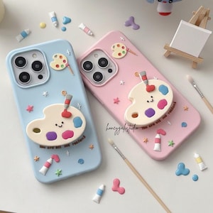 Puede incluir: Dos fundas para teléfono, una azul claro y otra rosa, decoradas con diseños temáticos de arte. Cada funda presenta una paleta de artista 3D con una cara sonriente, tubos de pintura y el texto "You are all my reasons".