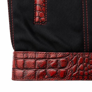 May include: Close-up of a black fabric bag with red stitching and a red crocodile-textured leather trim. The bag features a handle with matching red leather accents. The stitching is visible and adds a contrasting detail to the design.