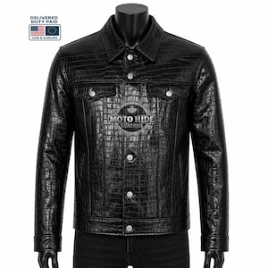 Può includere: Giacca in pelle nera con texture a effetto pelle di coccodrillo. Presenta chiusura con bottoni, due tasche sul petto e colletto classico. Esposta su manichino. Il logo "MOTO HIDE CUSTOMS" è visibile sul petto.