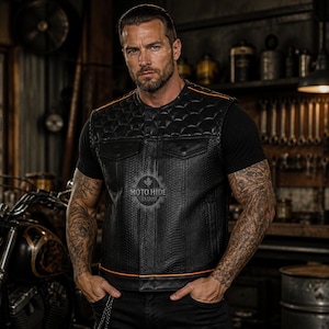 May include: Black leather vest with diamond stitching and orange trim. The vest has two front pockets and a Moto Hide logo. A motorcycle is visible in the background.