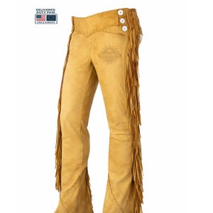 May include: Tan leather chaps with fringe detailing along the sides. The chaps feature a high waist with three white buttons and decorative stitching. A western-inspired garment.