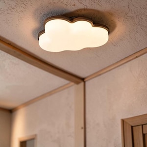 May include: A cloud-shaped ceiling light fixture. The light has a white, glowing cloud design with a brown trim. The fixture is mounted on a light-colored ceiling, with wooden beams and trim visible in the corners of the room.