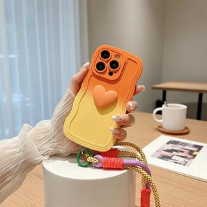 Könnte beinhalten: Orange-gelbes Farbverlauf-Handycase mit herzförmigem Akzent. Das Case hat ein wellenförmiges Kantendesign und ein mehrfarbiges Schlüsselband, das mit einem grünen Ring befestigt ist. Die Handyhülle dient zum Schutz des Telefons.
