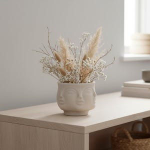 Könnte beinhalten: Eine beige Keramikvase mit drei Gesichtern, gefüllt mit getrockneten Blumen und Gräsern. Die Anordnung umfasst weißen Schleierkraut, beiges Pampasgras und andere getrocknete Stiele. Die Vase steht auf einer hellen Holzoberfläche.