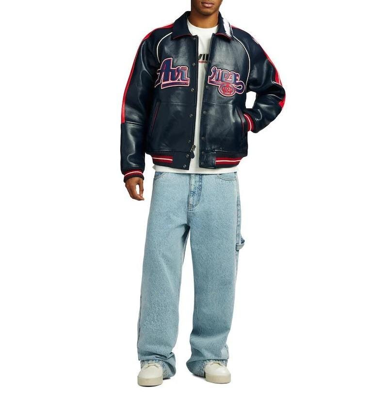 May include: A navy blue leather jacket with red and white accents, featuring embroidered text on the chest. The jacket is paired with light blue denim jeans and white sneakers. The overall style is casual and streetwear-inspired.